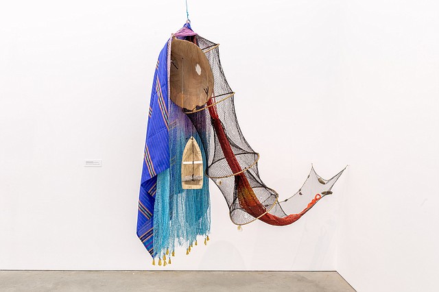Flying Massachusett, 2020. By Carolina Caycedo. Red de pesca artesanal teñida a mano, trampa de pesca artesanal con flotadores, hamaca artesanal teñida a mano, textiles tradicionales de la península de Zamboanga en Filipinas, sartén de oro de madera tallada a mano, barco de madera tallado a mano, concha blanca, aguja de madera, guijarros y piedras recogidas del puerto de Boston, Blue Hills Reservation y Neponset River, hilo de algodón y paracord, 93 x 166 x 23 pulgadas (236.2 x 421.6 x 58.4 cm). Cortesía del artista y de Commonwealth and Council, Los Ángeles.